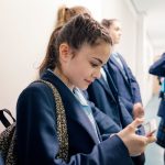 A group of students waiting in line in a school corridor, one is checking her phone